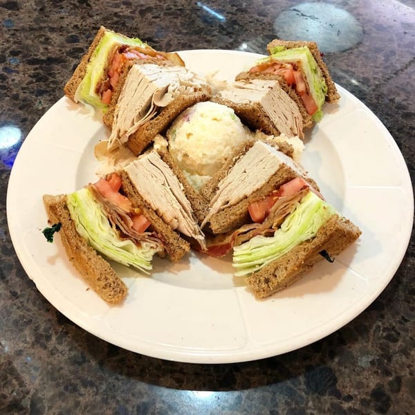 A Lighthouse Diner club sandwich, cut into quarters, with a scoop of potato salad on a white plate.