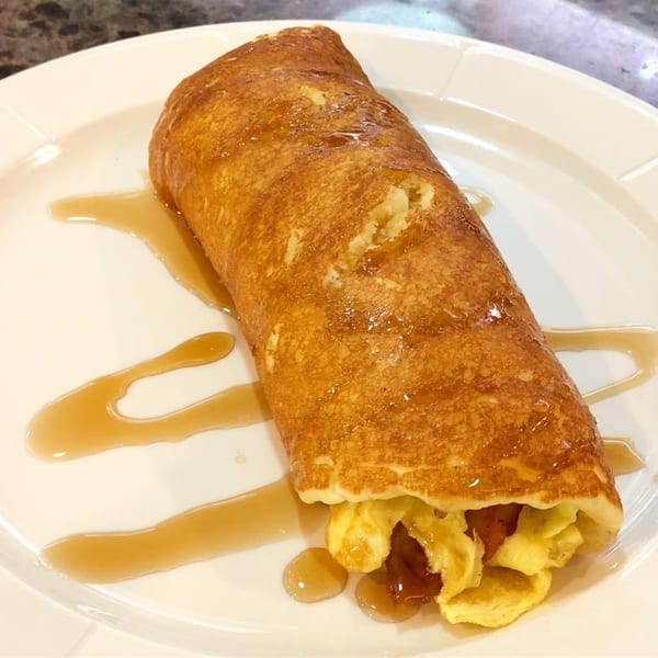 A golden-brown pancake roll filled with scrambled eggs and drizzled with maple syrup on a white plate.
